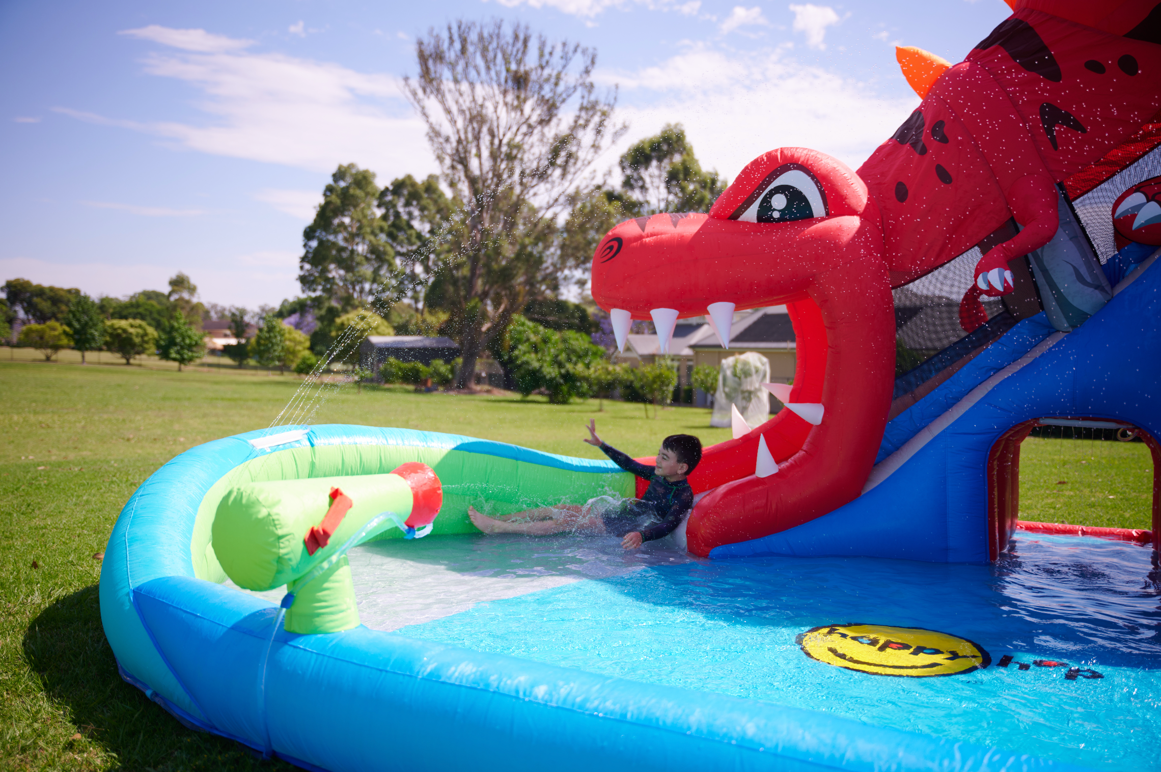 A children's inflatable bouncy castle with a dinosaur-themed slide and splash pool, set up in a grassy backyard with a child sliding down the red dinosaur mouth onto a blue water pool.